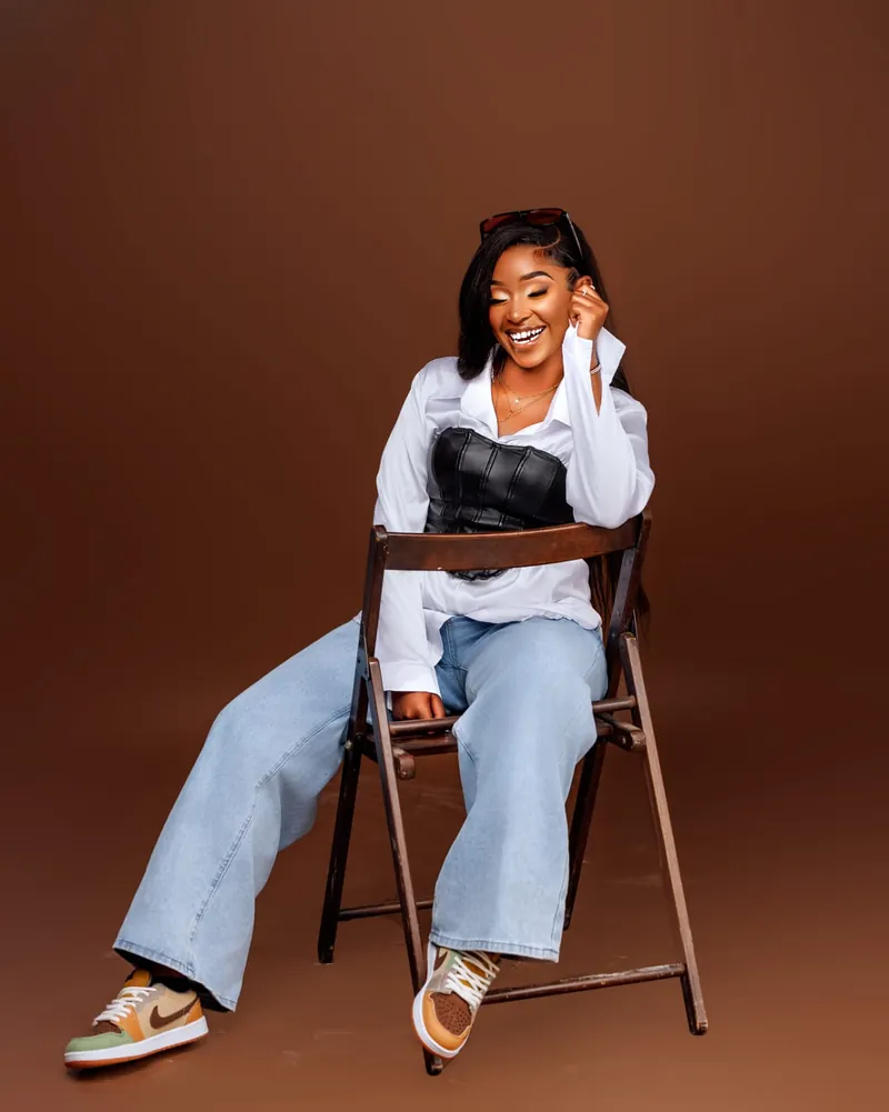 Wanja laughing on a wooden chair with one hand raised to her face, sunglasses pushed up in her hair